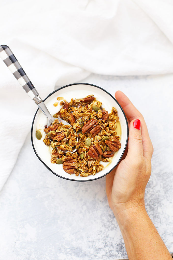 Hand holding a bowl of Maple Pumpkin Pecan Granola with Yogurt and a Black and White Gingham Spoon