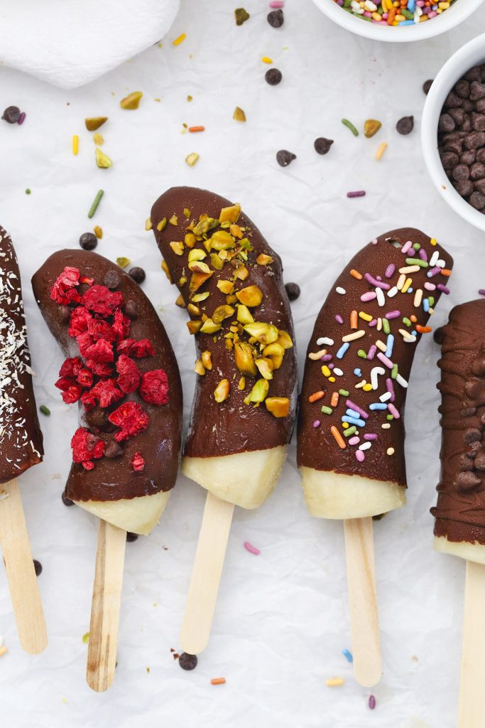 Close-up view of Frozen Chocolate Covered Bananas with different toppings on a piece of parchment paper