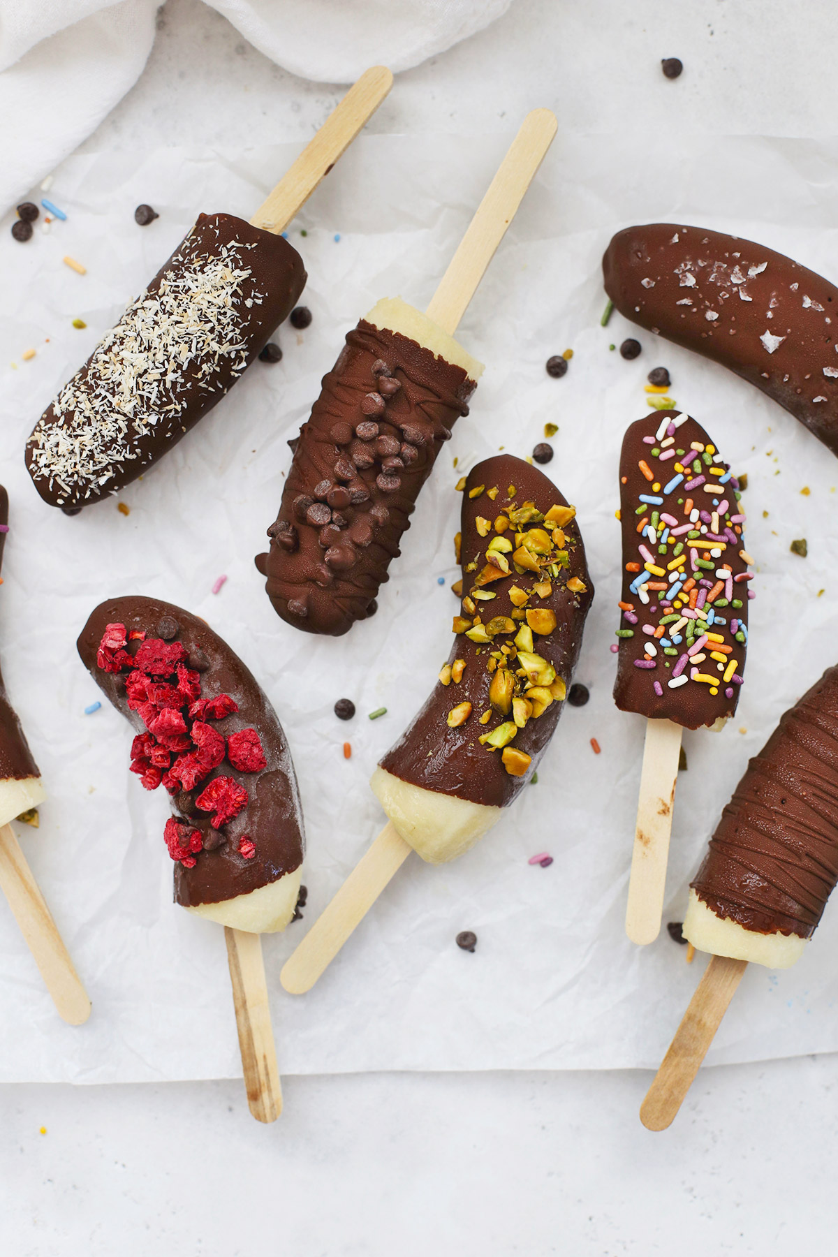 Frozen Chocolate Covered Bananas with different toppings scattered on a piece of parchment paper