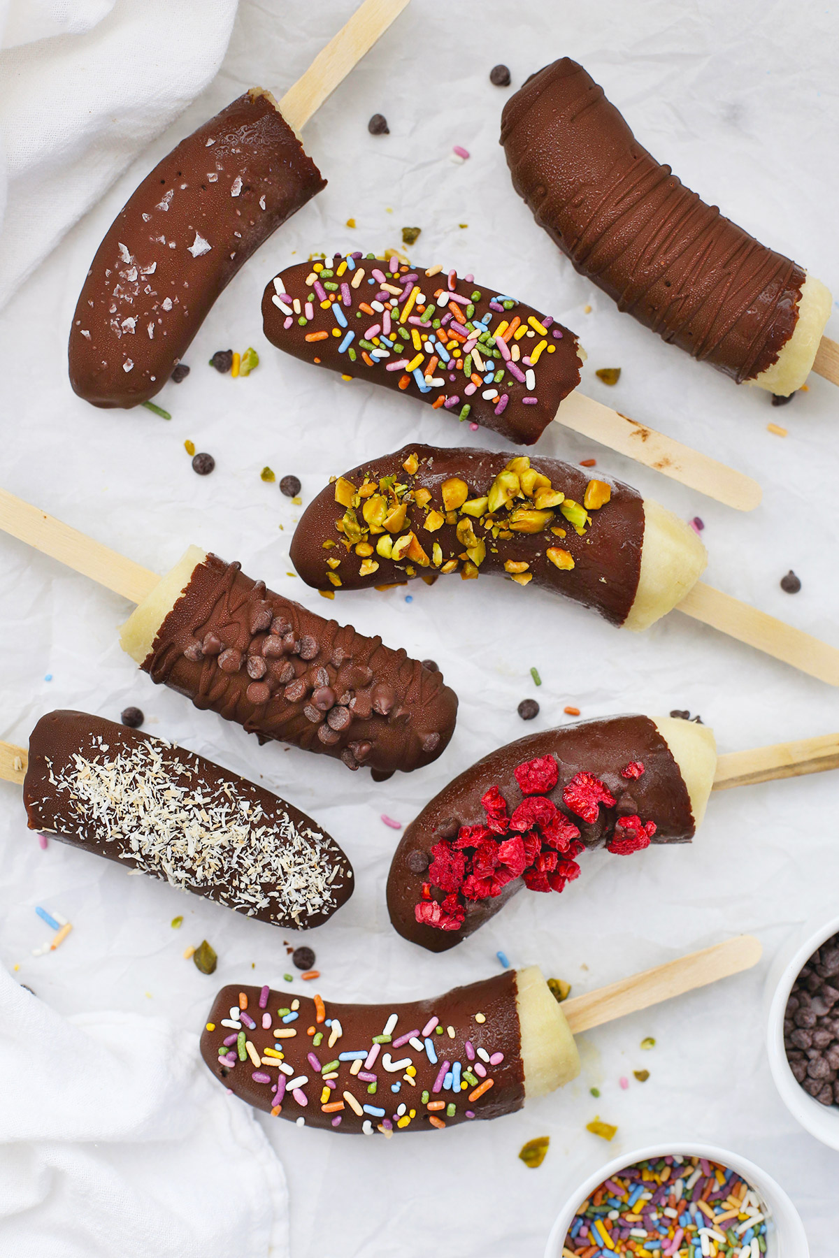 Frozen Chocolate Covered Bananas with different toppings scattered on a piece of parchment paper