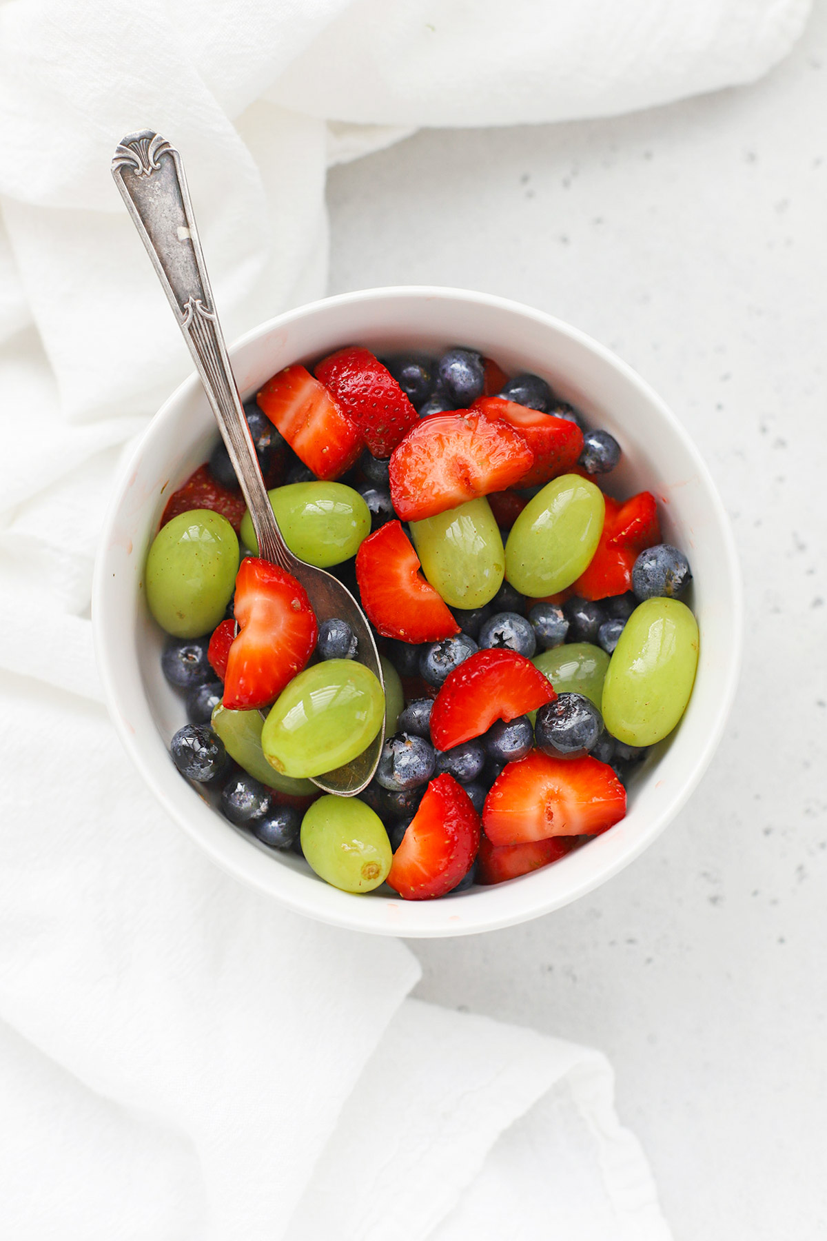 Close up overhead view of a small white bowl of honey lime fruit salad with strawberries, blueberries, and green grapes.