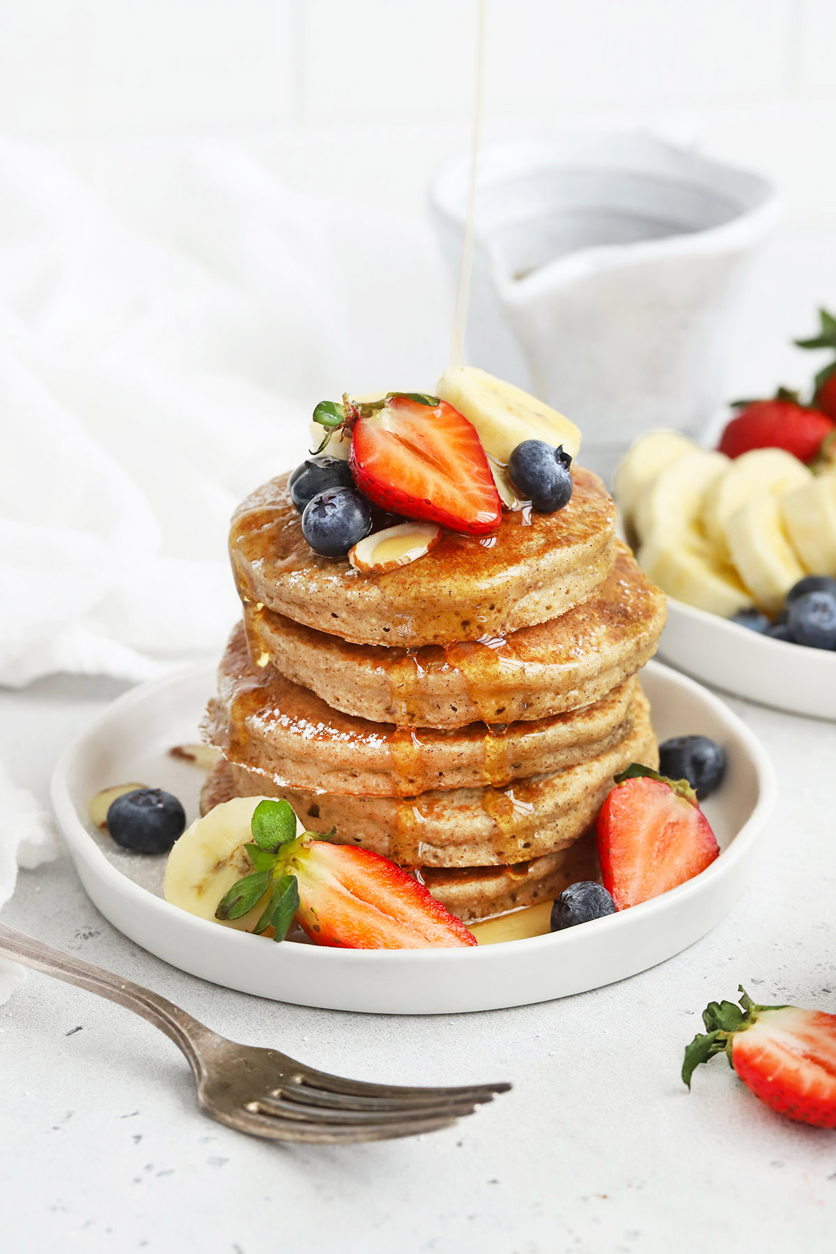 Front view of a stack of blender banana oatmeal pancakes topped with sliced bananas, fresh berries, and syrup.