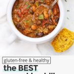 Overhead view of a white soup crock with healthy pumpkin chili inside and gluten-free pumpkin cornbread muffins on the side with text overlay that reads "gluten-free & healthy: the BEST EVER Pumpkin Chili: Easy + Healthy + Delicious!"