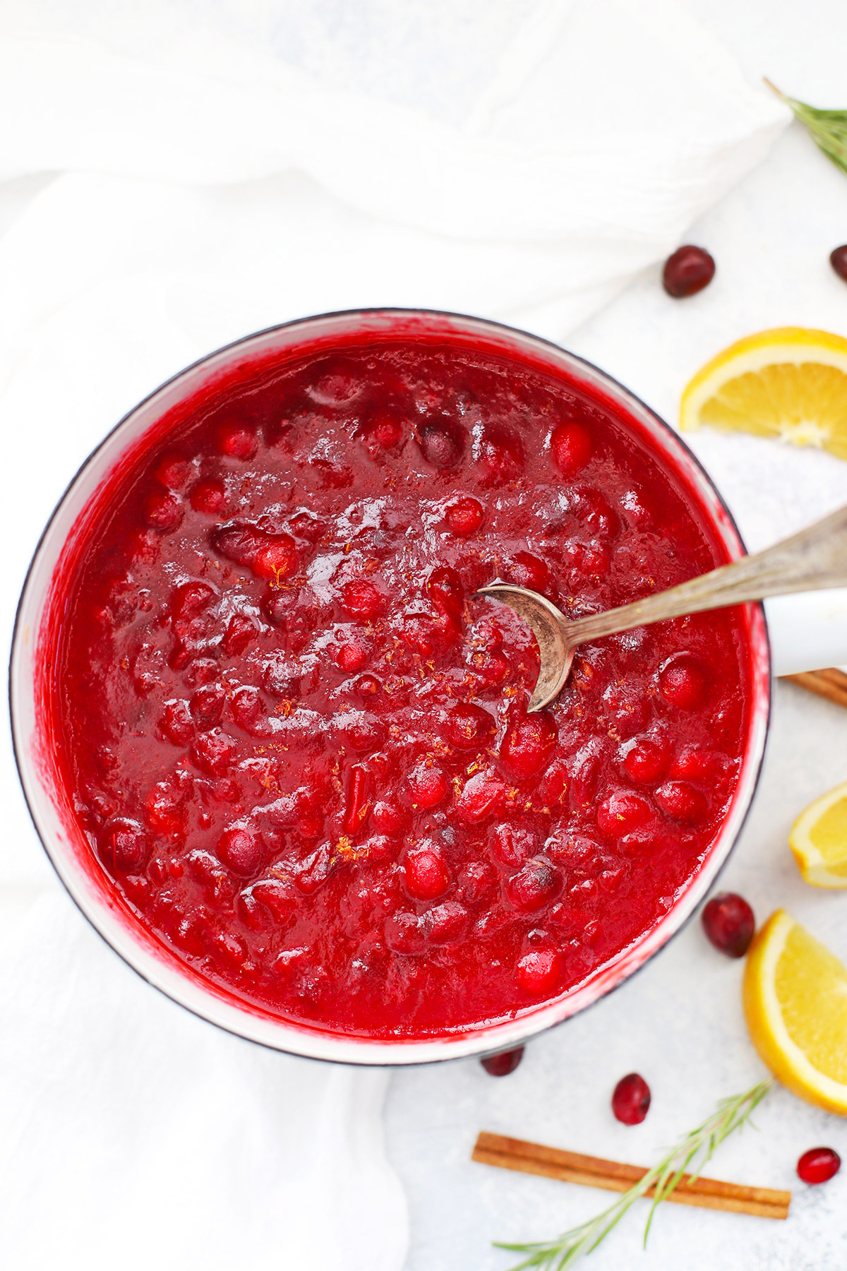 Overhead view of a white saucepan of paleo orange honey cranberry sauce with a spoon dipped in it. 