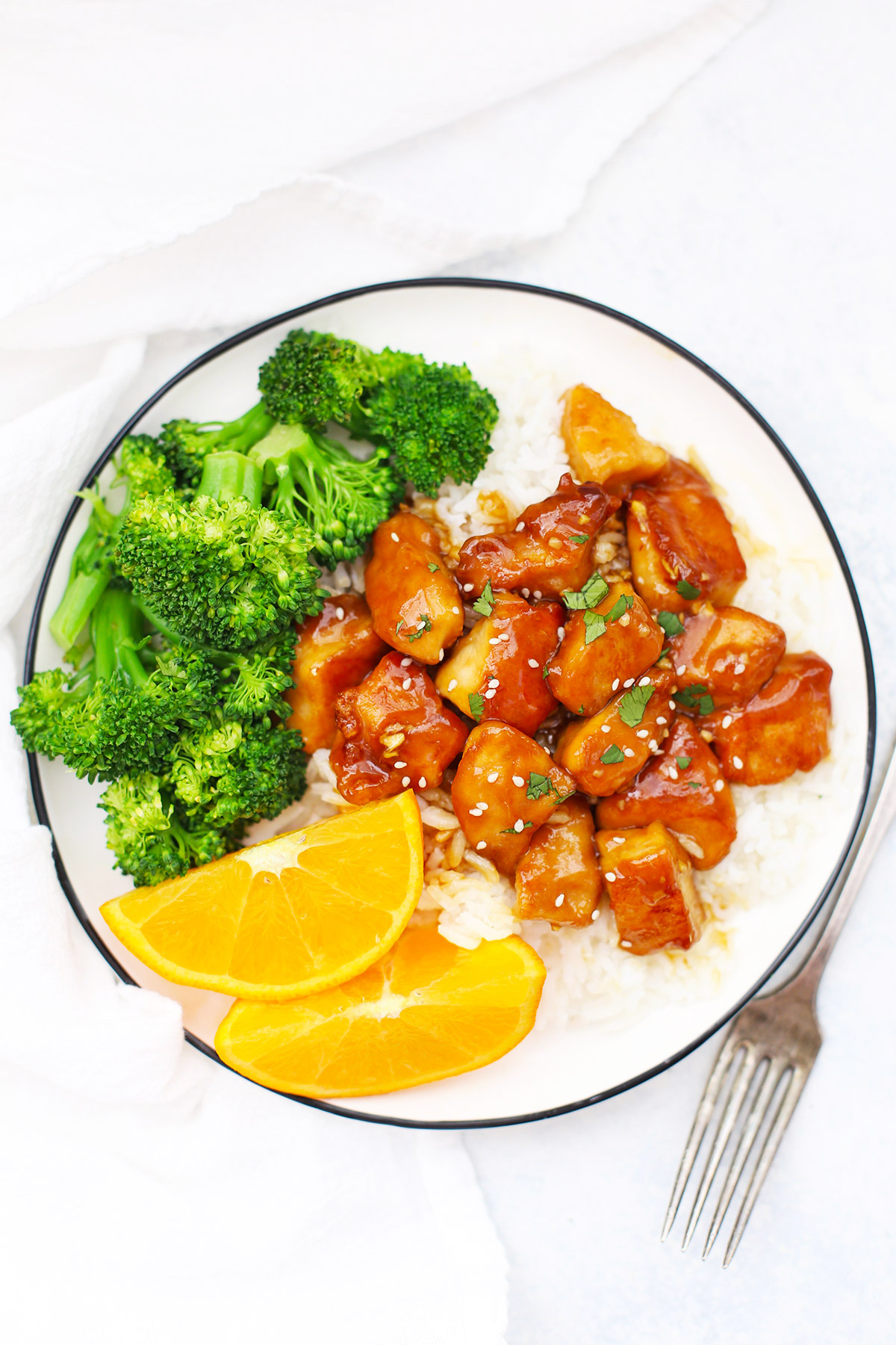 Overhead view of a plate with healthy orange chicken, rice, orange wedges and steamed broccoli on a white background