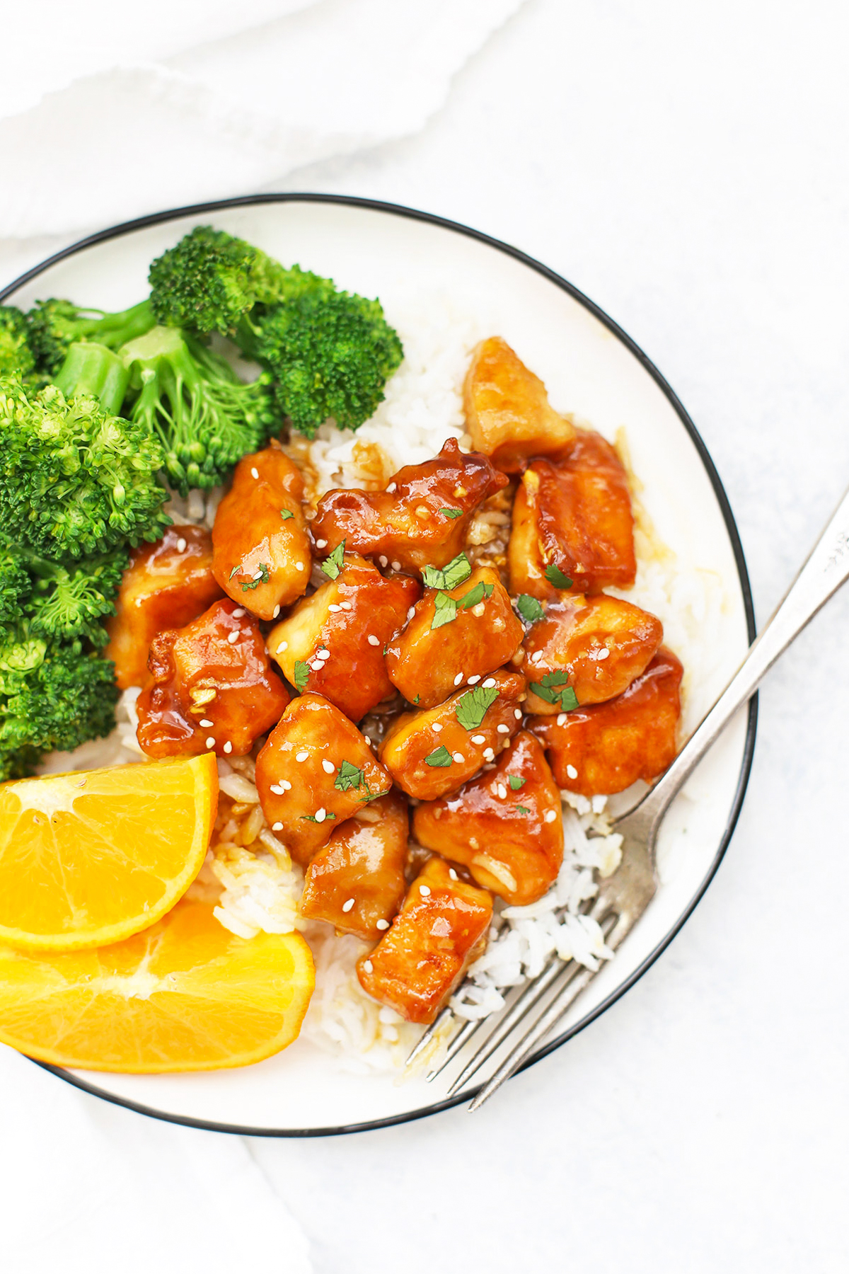Close up Overhead view of a plate with healthy orange chicken, rice, orange wedges and steamed broccoli on a white background