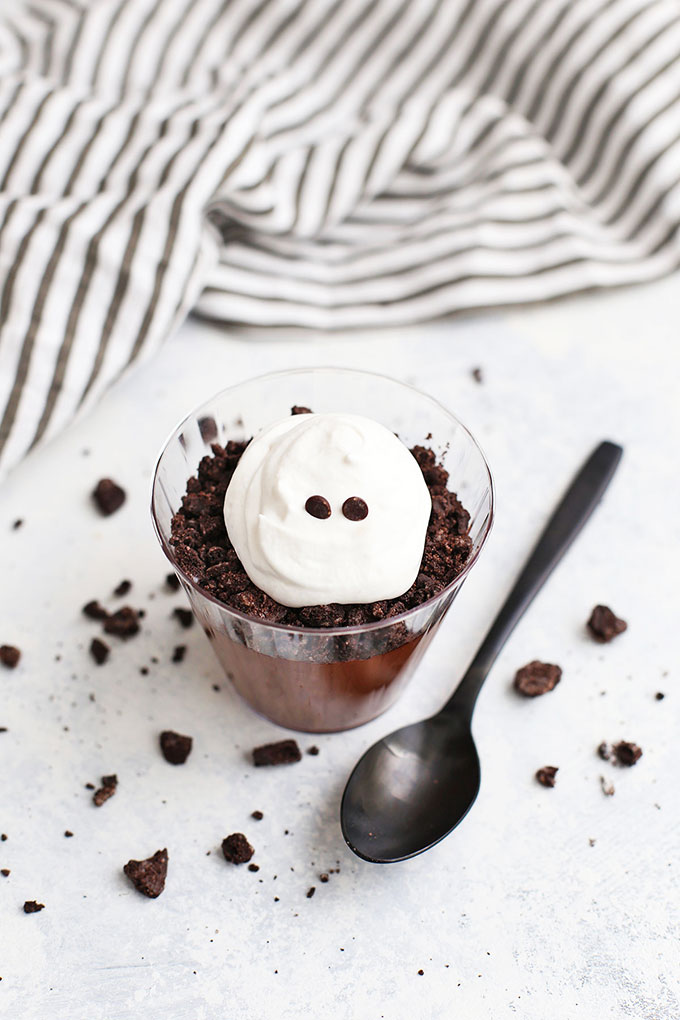 Whipped Cream Ghost with chocolate chip eyes in a dirt pudding cup with black spoon next to the cup. 