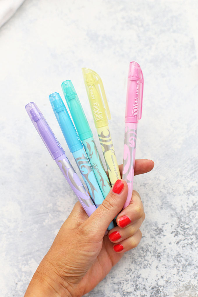 Hand holding erasable highlighters in lilac, aqua, mint, gold, and blush. 