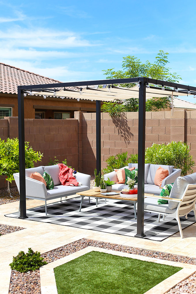 Outdoor living area with pergola, grey outdoor sofas, grey outdoor chairs, black and white gingham rug and pink and green accessories.