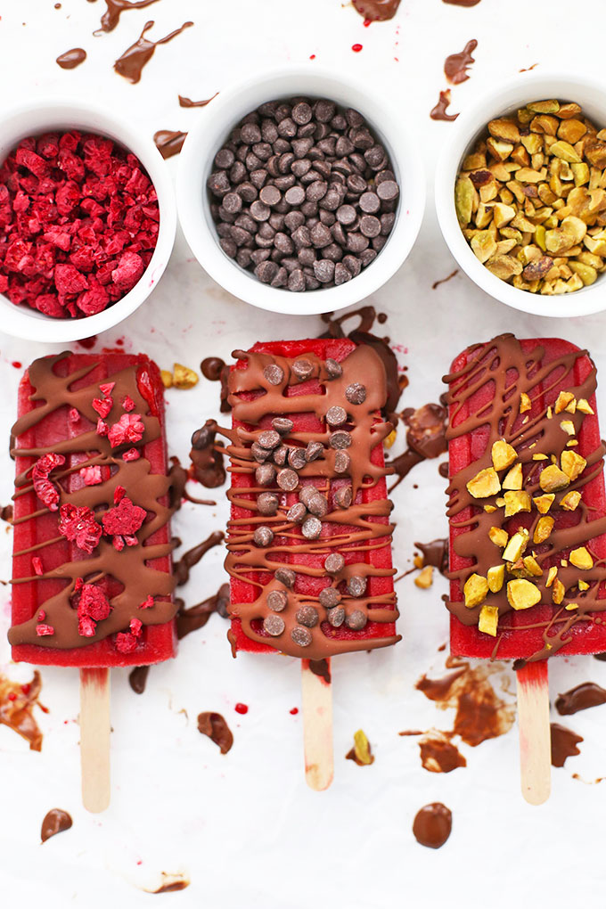 Paleo & Vegan Raspberry Sorbet Popsicles on a baking sheet, drizzled with chocolate shell and sprinkled with mini chocolate chips, chopped pistachios, and freeze dried raspberries