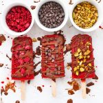 Paleo & Vegan Raspberry Sorbet Popsicles on a baking sheet, drizzled with chocolate shell and sprinkled with mini chocolate chips, chopped pistachios, and freeze dried raspberries