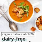 Overhead view of a bowl of dairy-free creamy tomato basil soup topped with gluten-free croutons and basil chiffonade with text overlay that reads "vegan + paleo + whole30 dairy-free tomato basil soup: warm + cozy + velvety smooth"