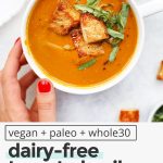 Overhead view of a hand setting down a bowl of dairy-free creamy tomato basil soup topped with gluten-free croutons and basil chiffonade with text overlay that reads "vegan + paleo + whole30 dairy-free tomato basil soup: warm + cozy + velvety smooth"