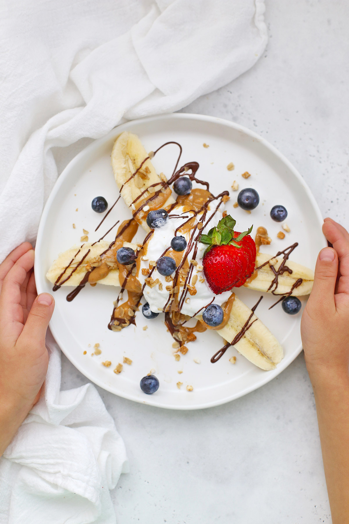 Hands holding a Healthy Banana Split with Yogurt, Peanut Butter, Chocolate, and berries on a white plate