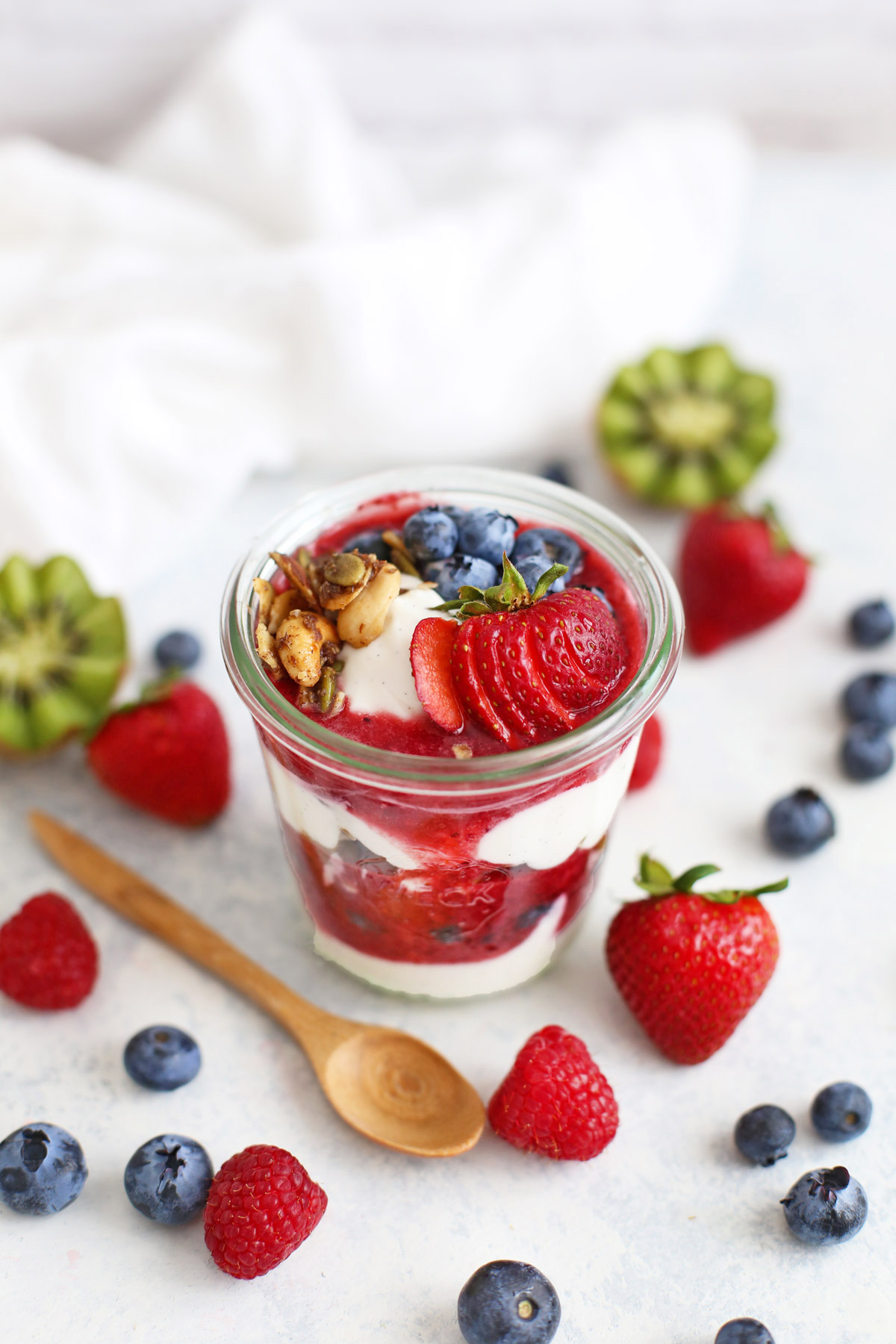 Paleo parfait in a jar with coconut milk yogurt, strawberry puree, and grain-free granola, with fresh fruit scattered in the background.