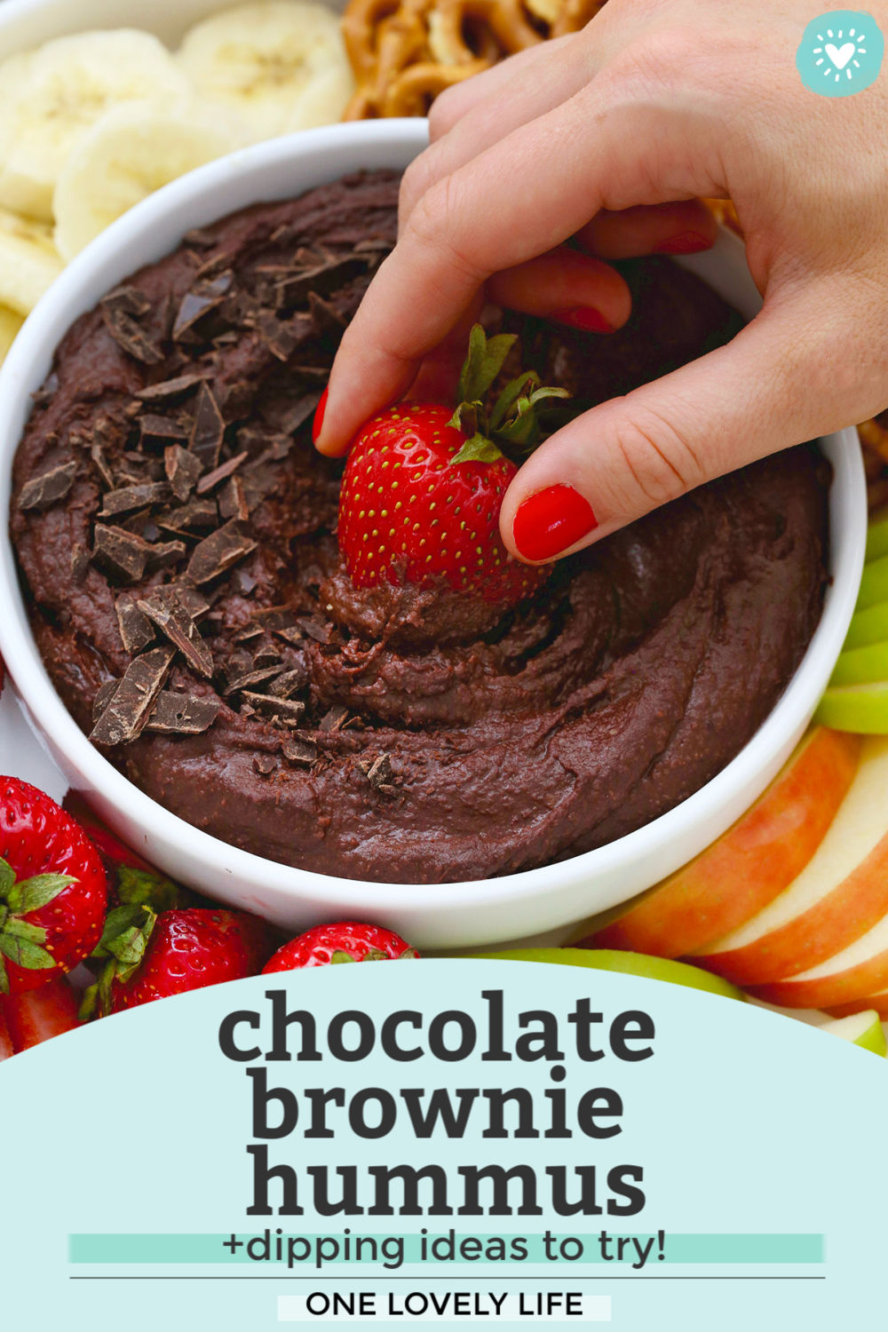 A strawberry being dipped into chocolate brownie hummus on a fruit plate with text overlay that reads "Chocolate brownie hummus +dipping ideas to try! One Lovely Life"