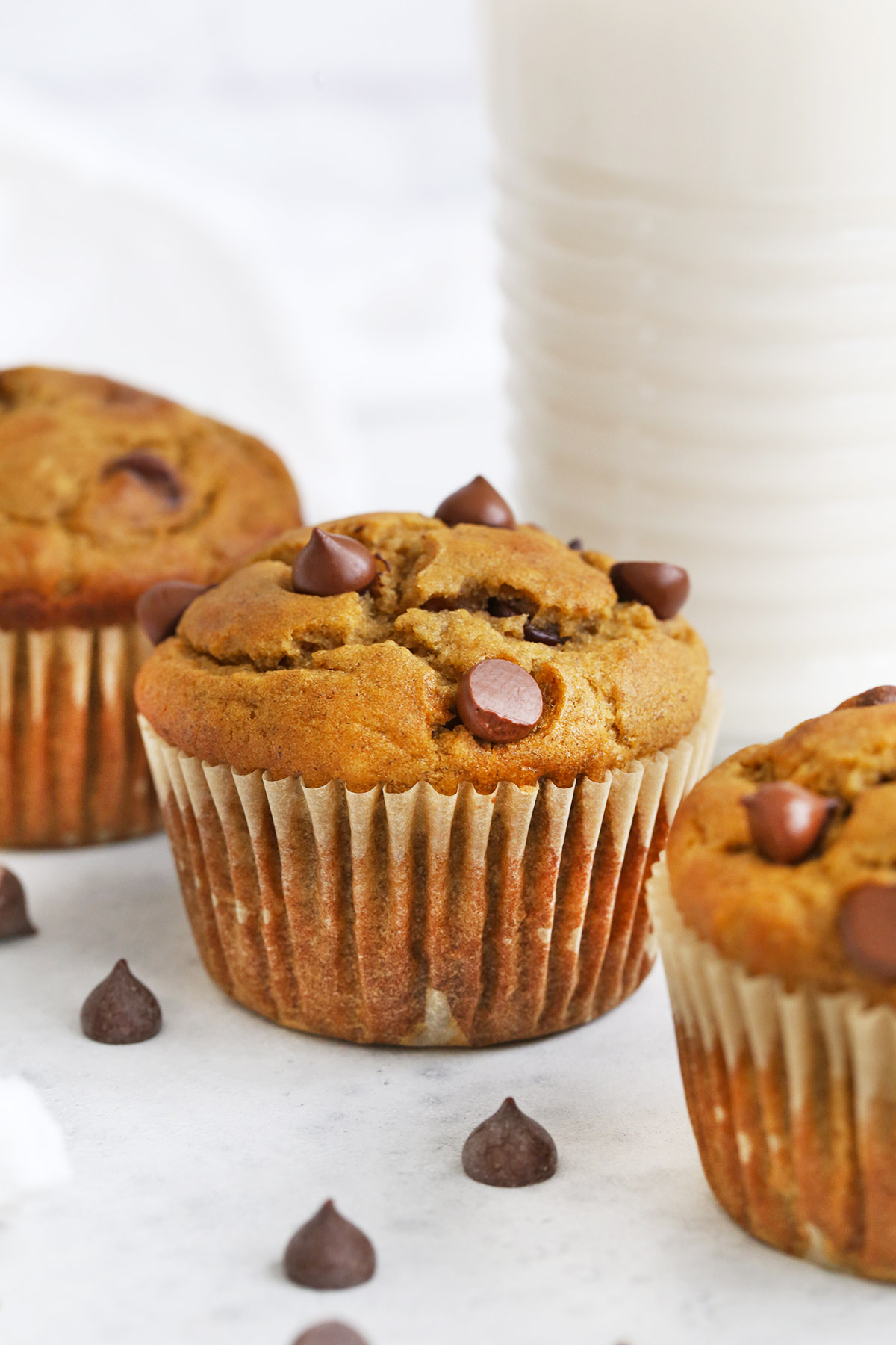 Three gluten-free chocolate chip banana muffins on a white background with a glass of almond milk