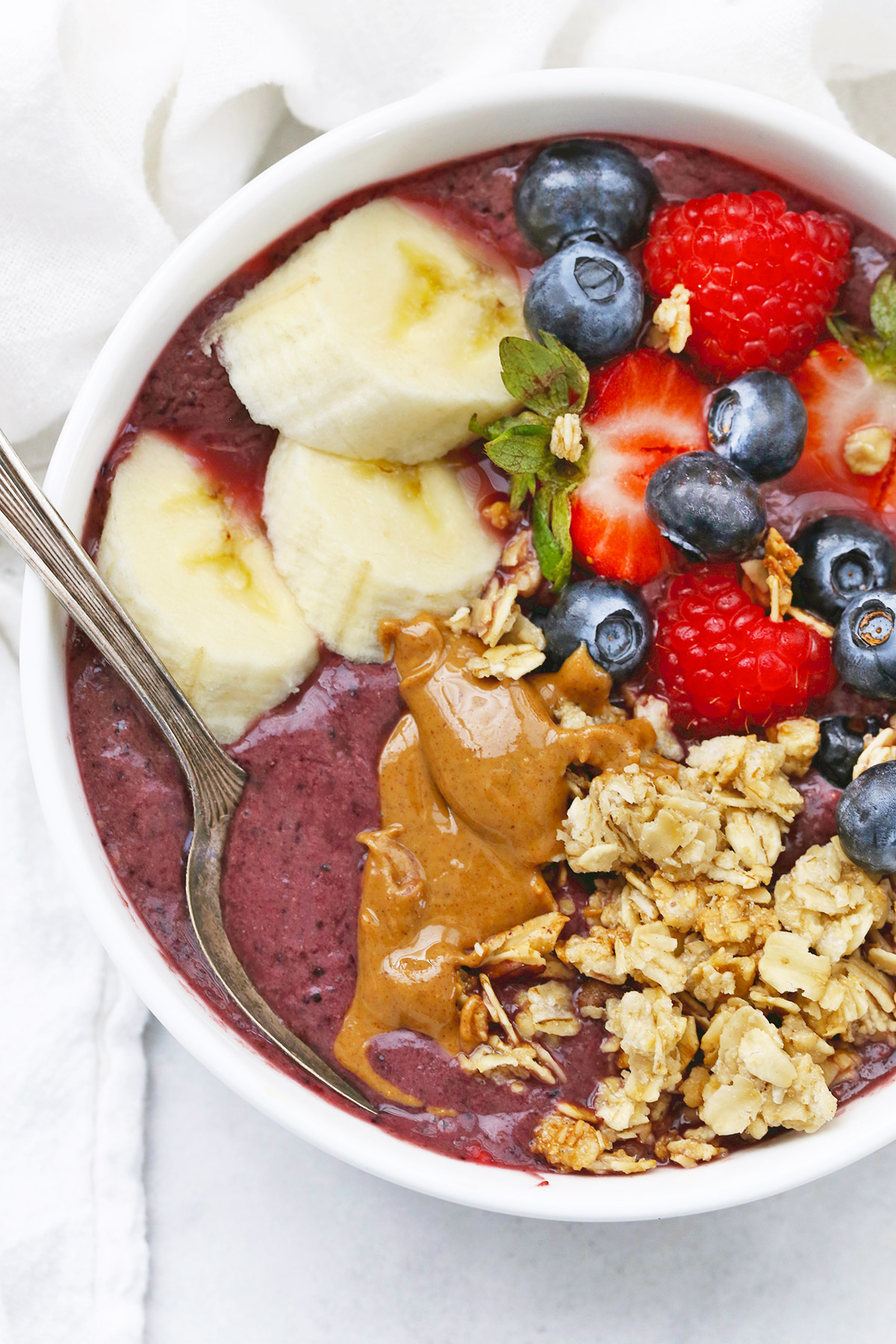 Close-up overhead view of a peanut butter acai bowl topped with fresh berries, peanut butter, granola, and banana slices on a white background