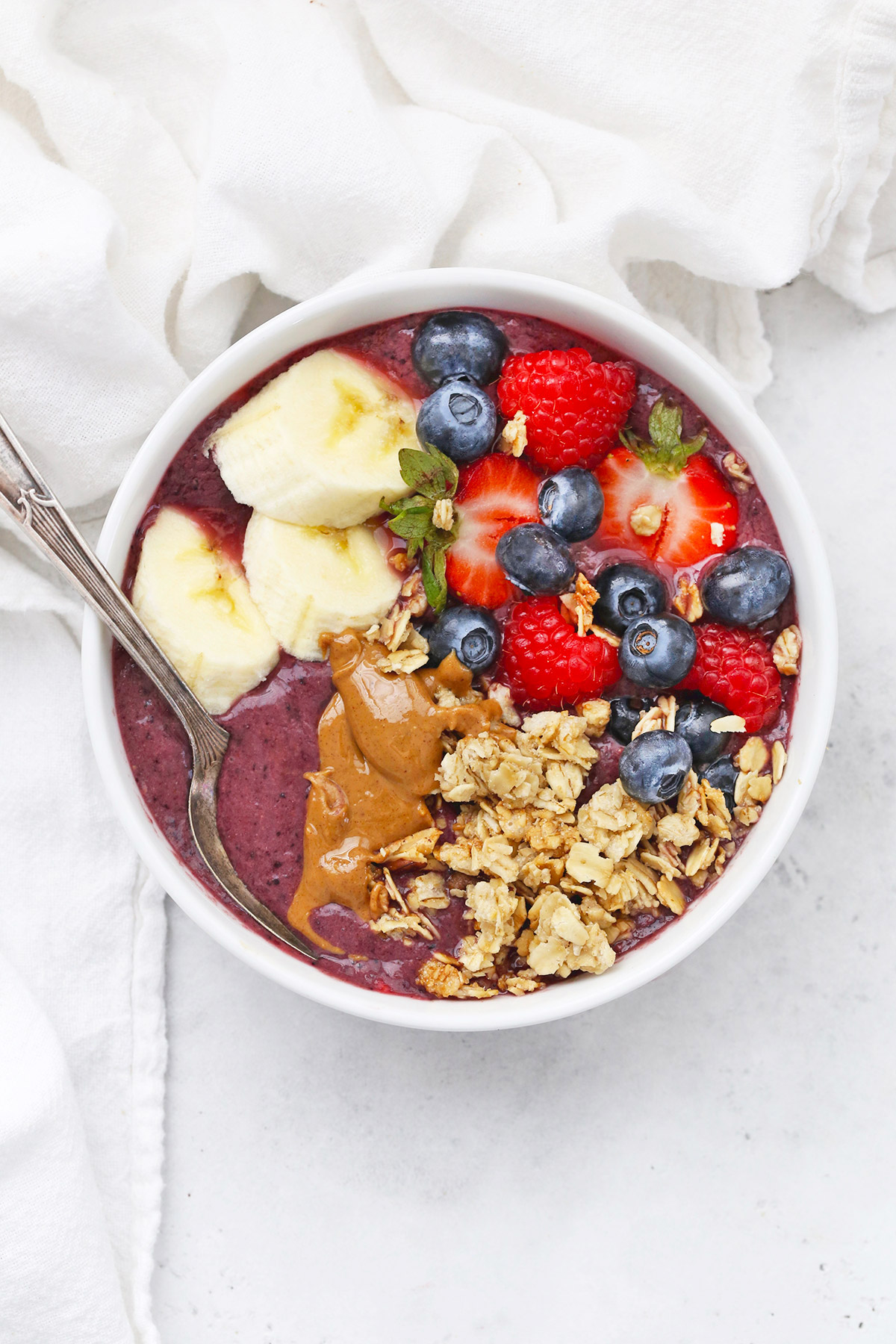 Overhead view of a peanut butter acai bowl topped with fresh berries, peanut butter, granola, and banana slices on a white background