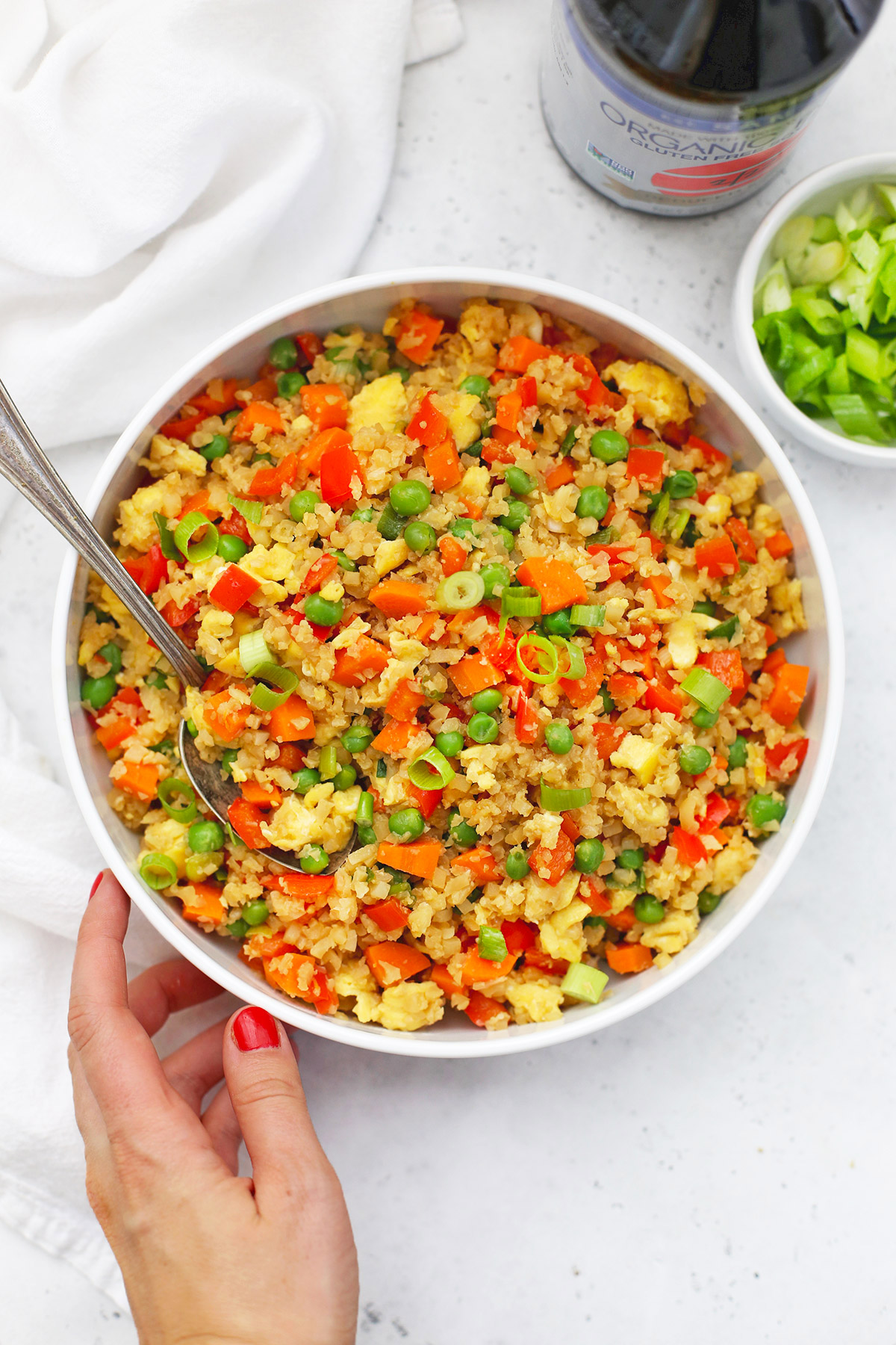 One hand reaching for A large white bowl of cauliflower fried rice on a white background. 