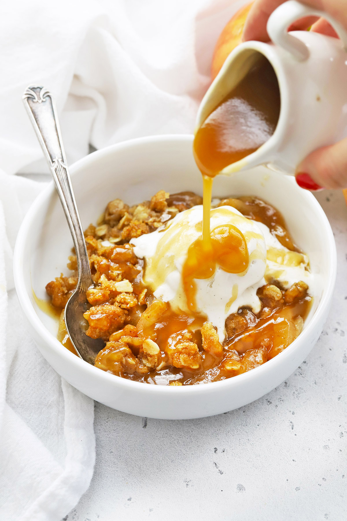 Pouring cider caramel sauce over a bowl of gluten-free apple crisp with a scoop of vanilla ice cream
