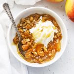 A white bowl of gluten-free apple crisp topped with vanilla ice cream and cider caramel sauce