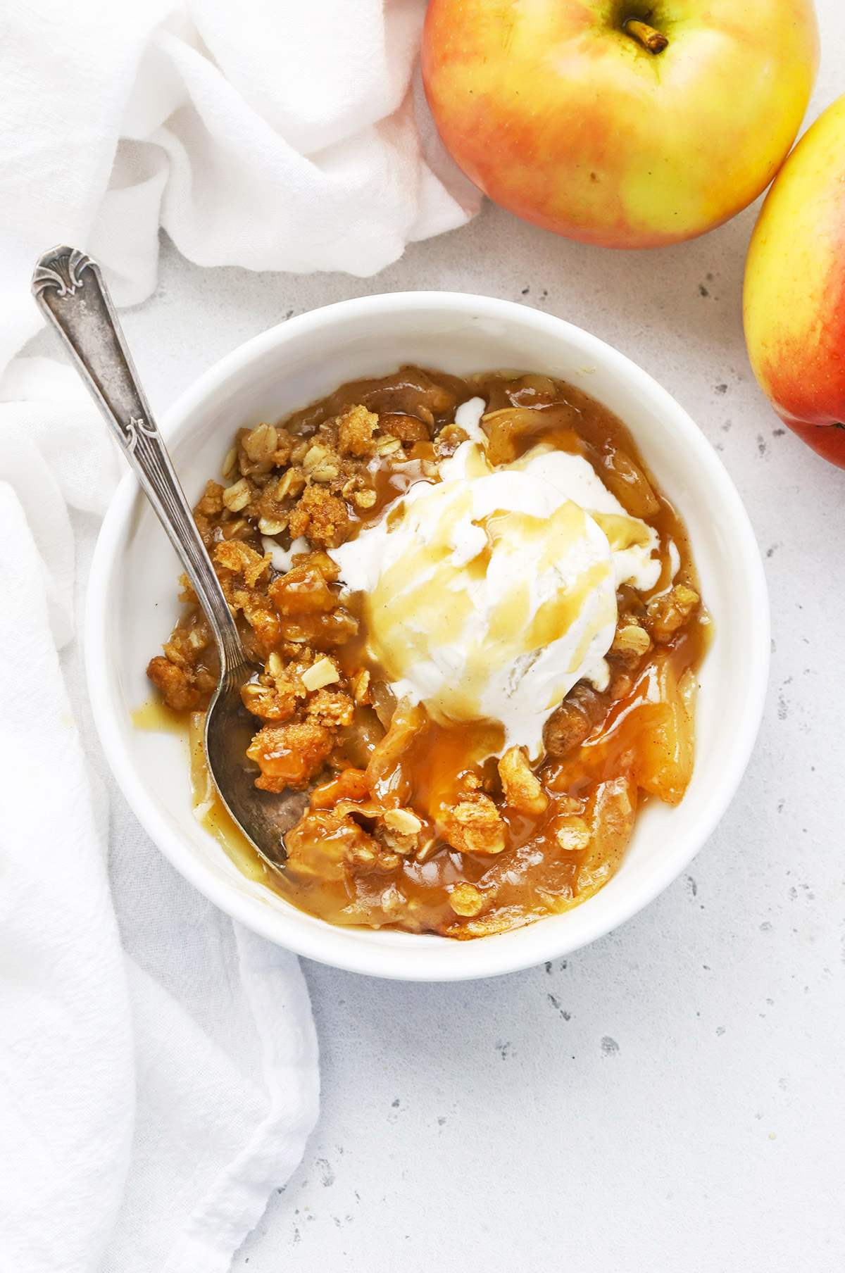 A white bowl of gluten-free apple crisp topped with vanilla ice cream and cider caramel sauce