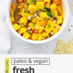 Overhead view of Mango Salsa in a white bowl with tortilla chips on the side with text overlay that reads "paleo & vegan fresh mango salsa: sweet + savory + spicy"