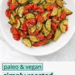Overhead view of a bowl of simply roasted zucchini and tomatoes with text overlay that reads "vegan + paleo simply roasted zucchini & tomatoes: the seasoning is amazing!"