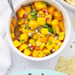 Overhead view of Mango Salsa in a white bowl with tortilla chips on the side with text overlay that reads "Fresh mango salsa. Vegan + paleo + crazy delicious!"