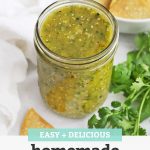 Front view of a jar of homemade salsa verde (green salsa) with fresh cilantro and tortilla chips in the background with text overlay that reads "Easy + Delicious Homemade Salsa Verde"