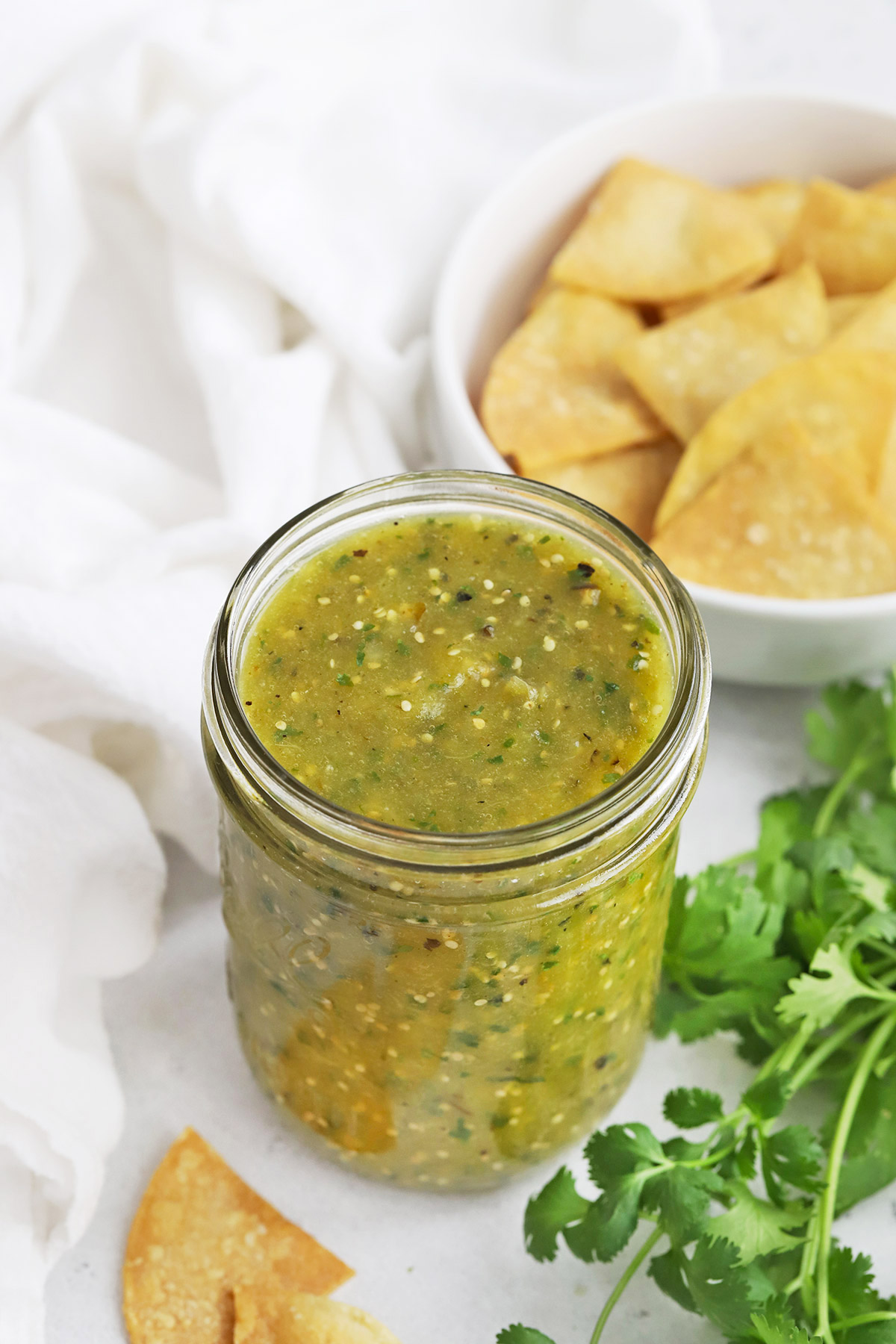 Front view of a jar of homemade salsa verde (green salsa) with fresh cilantro and tortilla chips in the background