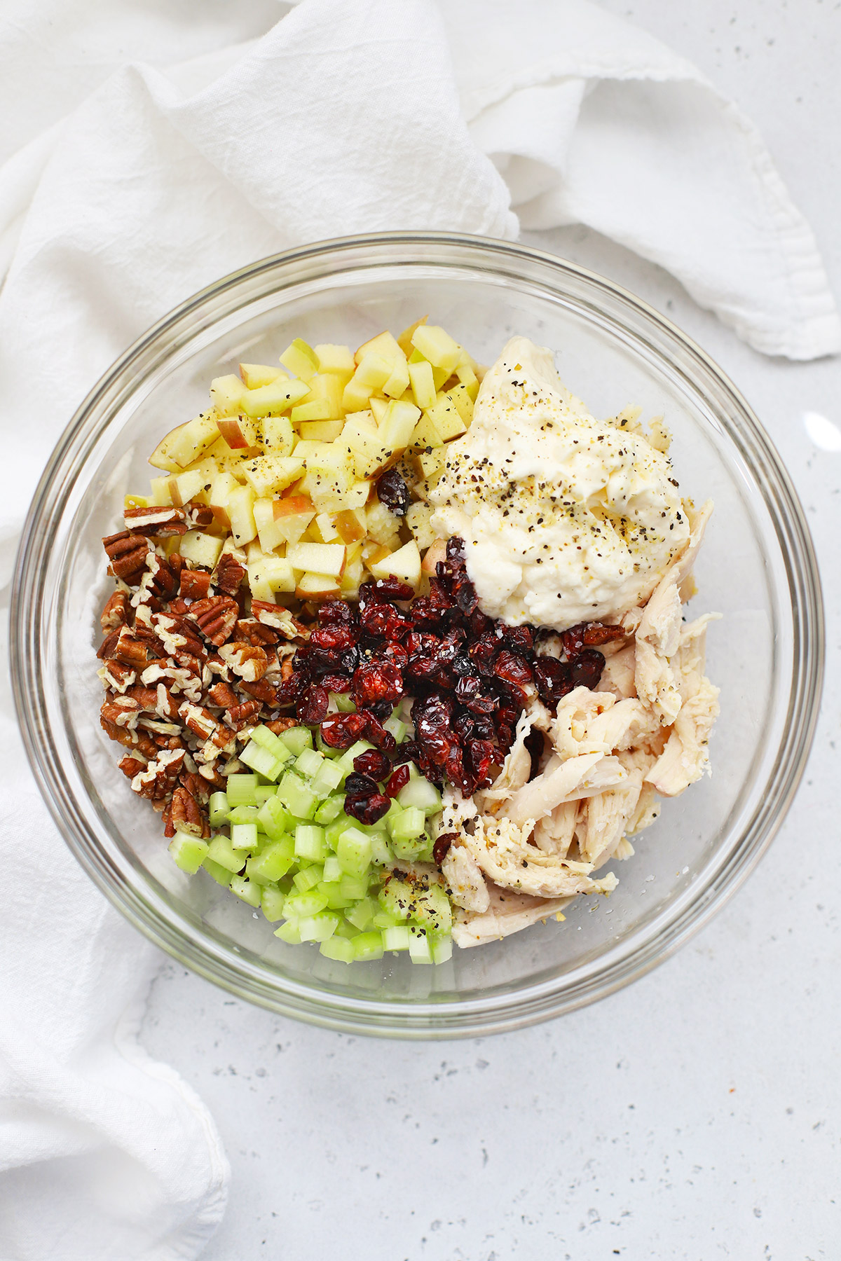 Overhead view of a glass bowl of ingredients for Apple Cranberry Chicken Salad--shredded chicken, diced apples, celery, dried cranberries, mayo, and seasonings. 