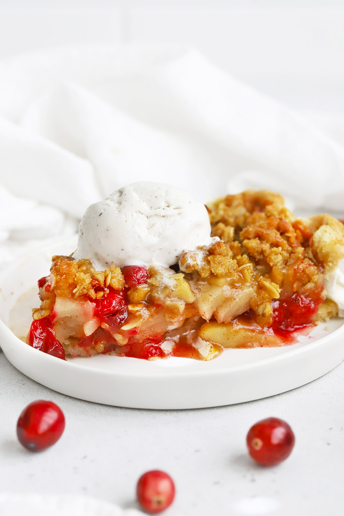 Front view of a slice of apple cranberry crumble pie topped with vanilla ice cream on a white plate