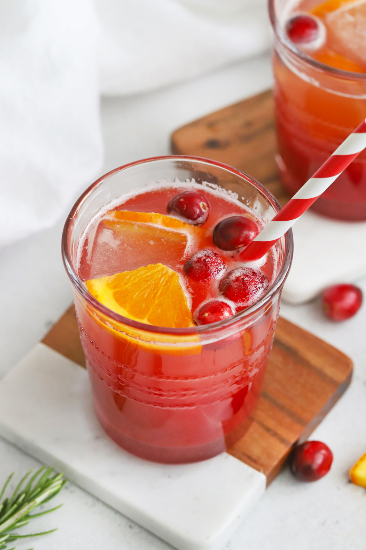 Front view of two Cranberry Cider Mocktails garnished with ice, orange wedges and fresh cranberries on marble and wood coasters