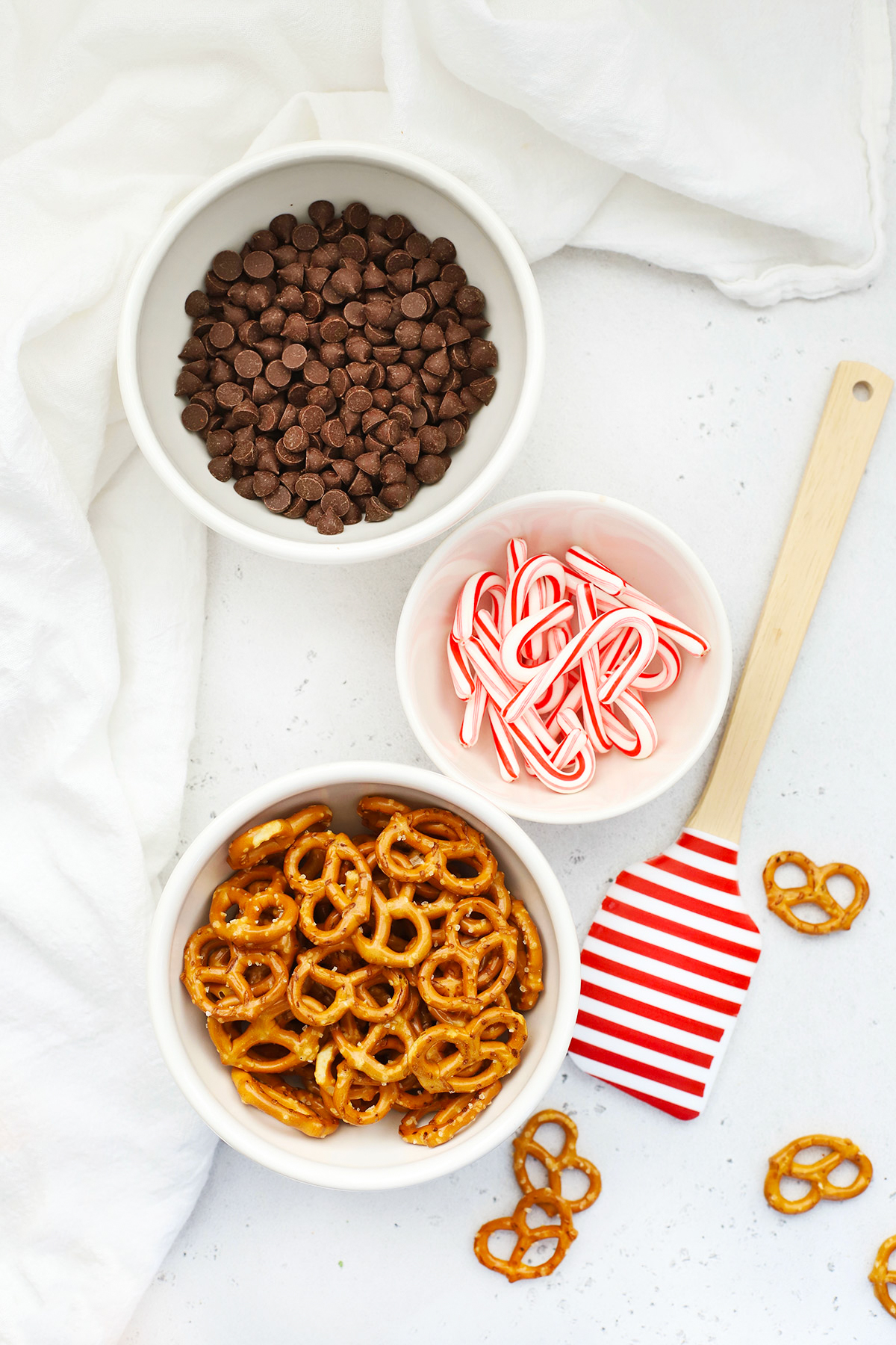 Overhead view of 3 bowls of ingredients for gluten-free chocolate peppermint pretzels--vegan chocolate chips, mini candy canes, and gluten-free pretzels.