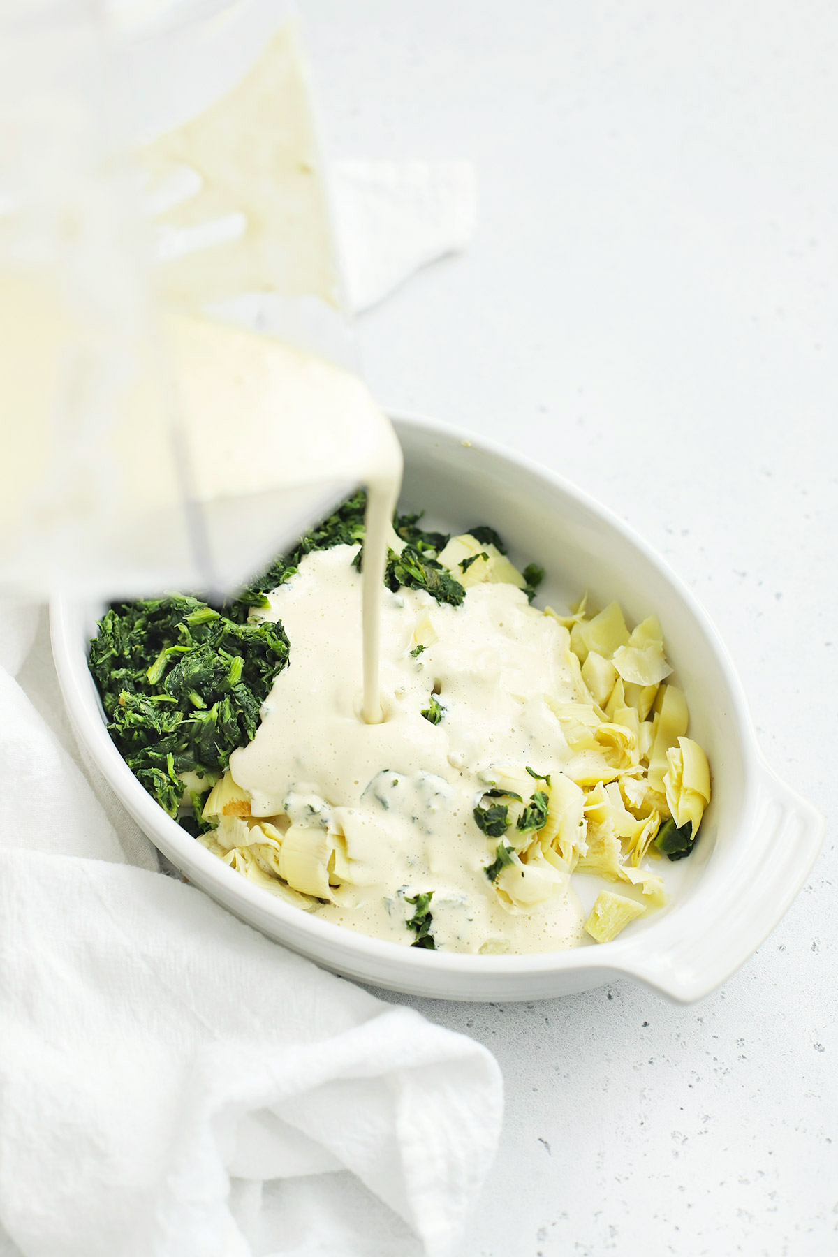 Pouring creamy cashew sauce over spinach and artichokes to make vegan spinach artichoke dip