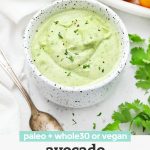 Front view of a bowl of paleo or vegan avocado ranch with colorful veggies on a platter behind with text overlay that reads "Paleo + Whole30, or vegan avocado ranch dressing"