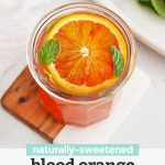 Front view of a blood orange mocktail garnished with fresh mint and blood oranges in the background with text overlay that reads "naturally-sweetened blood orange mocktail"