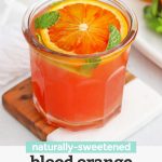 Front view of a blood orange mocktail garnished with fresh mint and blood oranges in the background with text overlay that reads "naturally-sweetened blood orange mocktail"