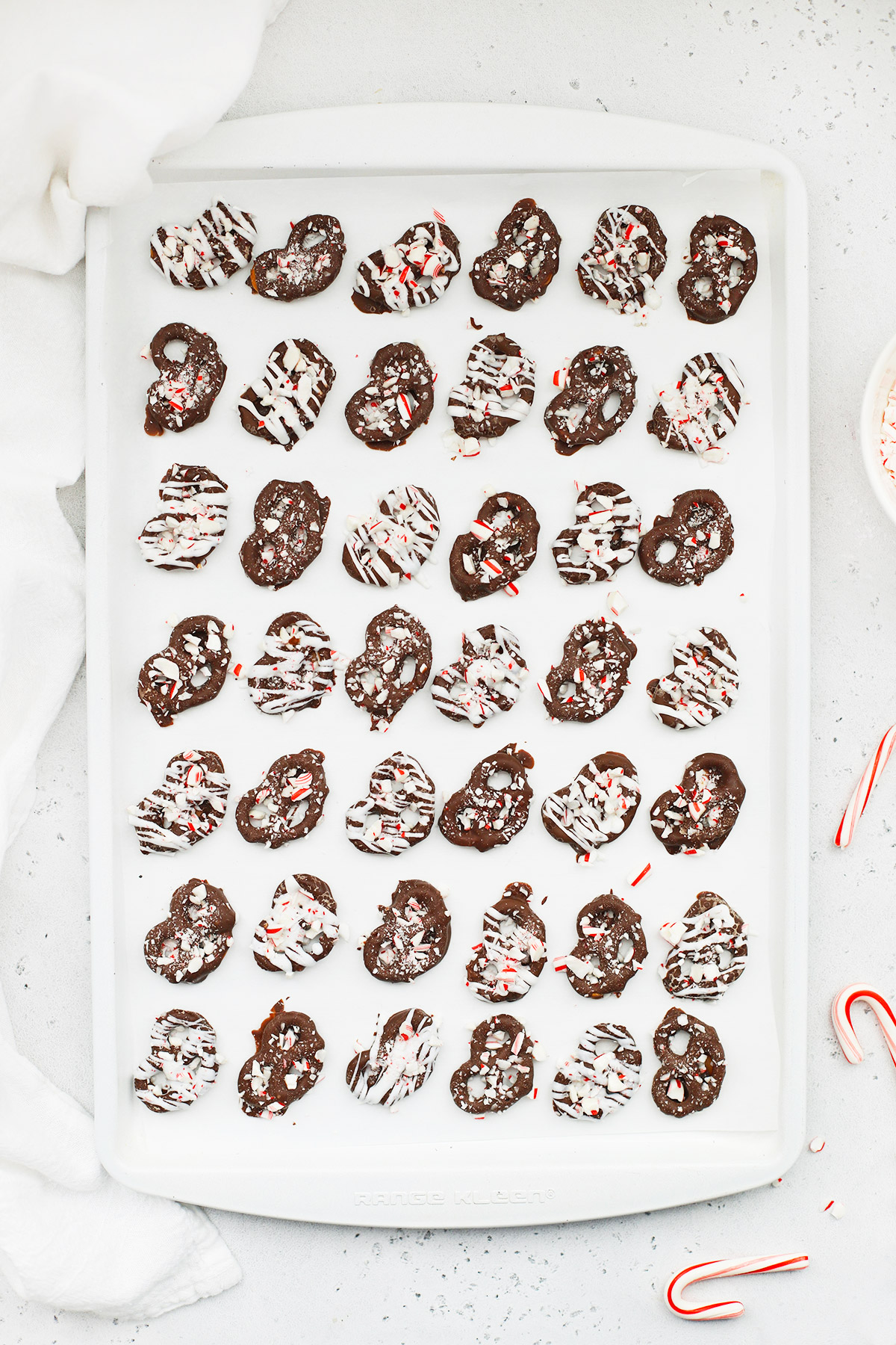 Overhead view of gluten-free chocolate peppermint pretzels covered with crushed candy canes cooling on a white baking sheet