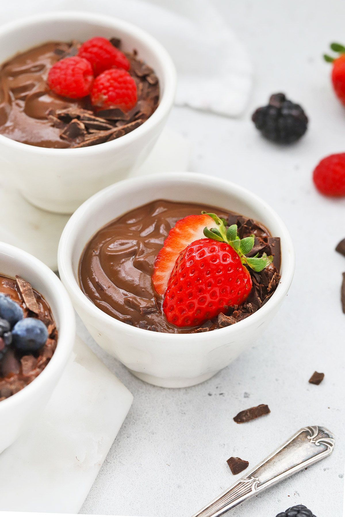 Close up front view of small white bowls of avocado chocolate mousse topped with fresh berries and shaved chocolate