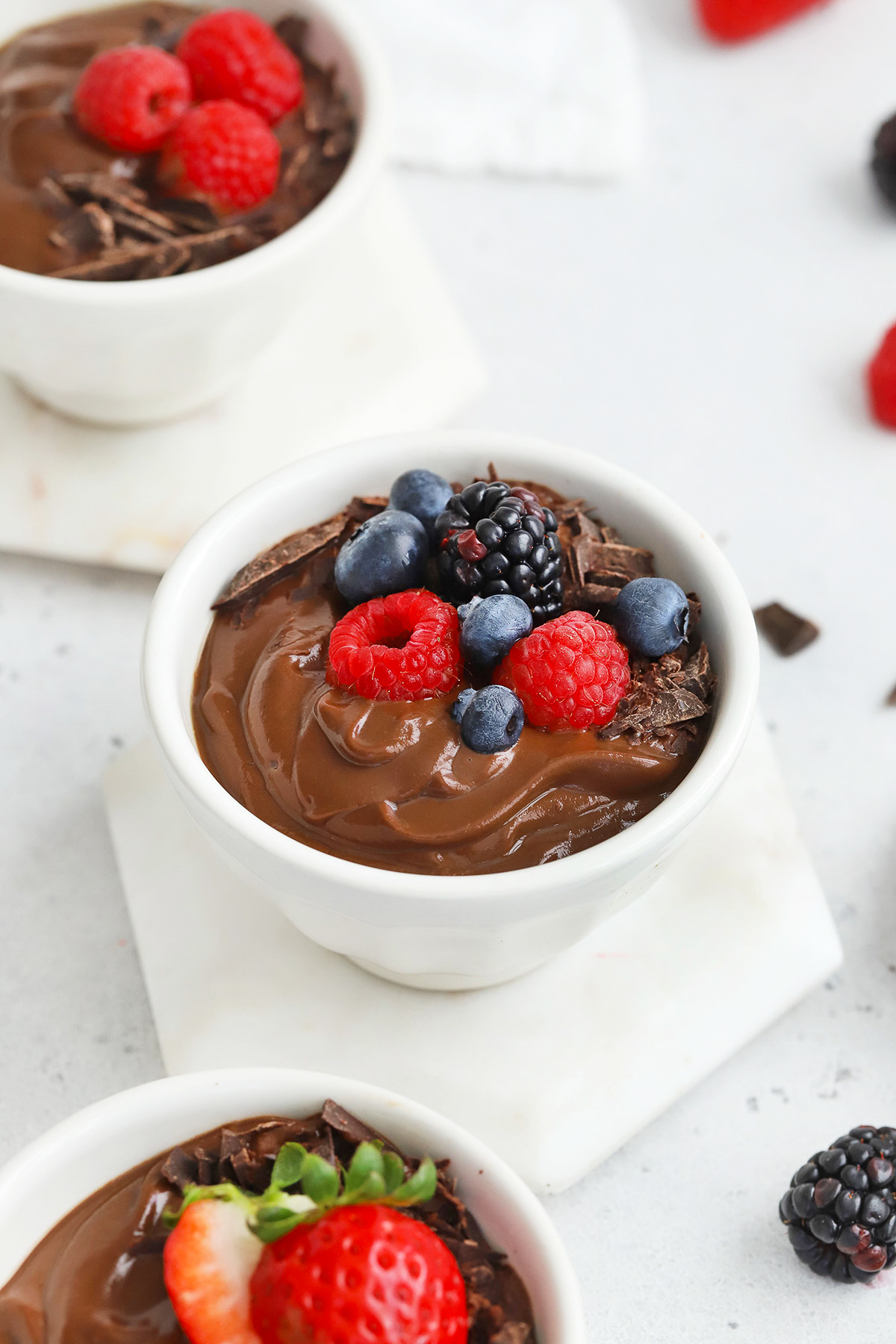 Close up front view of small white bowls of avocado chocolate mousse topped with fresh berries and shaved chocolate