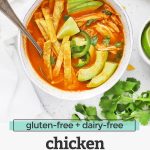 Overhead view of a white stoneware bowl of chicken tortilla soup topped with sliced avocado and crispy baked tortilla strips with text overlay that reads "gluten-free + dairy-free chicken tortilla soup. -One Lovely Life-"