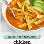Front view of a white stoneware bowl of chicken tortilla soup topped with sliced avocado and crispy baked tortilla strips with text overlay that reads "gluten-free + dairy-free chicken tortilla soup. -One Lovely Life-"