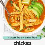 Overhead view of a white stoneware bowl of chicken tortilla soup topped with sliced avocado and crispy baked tortilla strips with text overlay that reads "gluten-free + dairy-free chicken tortilla soup. -One Lovely Life-"
