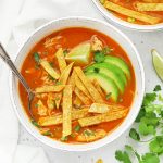 Overhead view of a white stoneware bowl of chicken tortilla soup topped with sliced avocado and crispy baked tortilla strips.