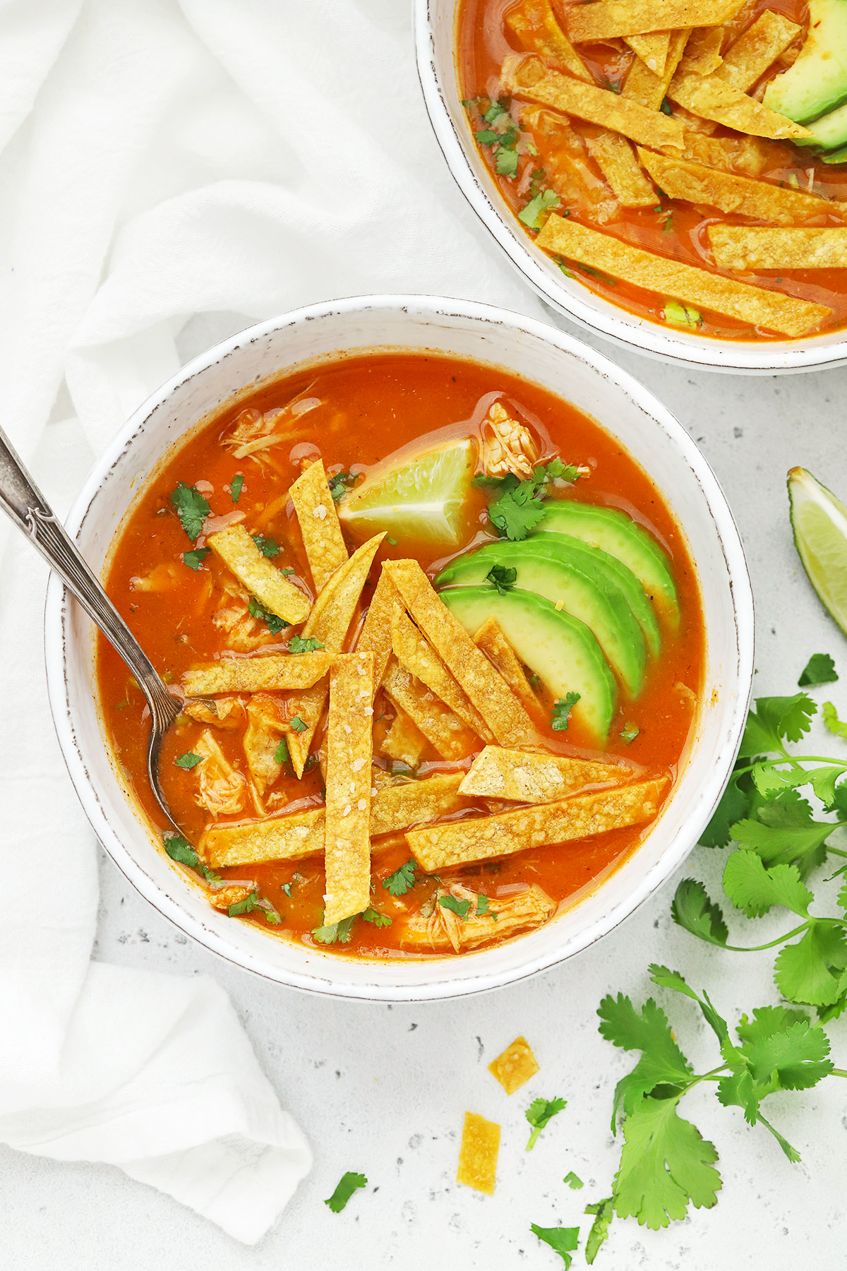 Overhead view of a white stoneware bowl of chicken tortilla soup topped with sliced avocado and crispy baked tortilla strips. 