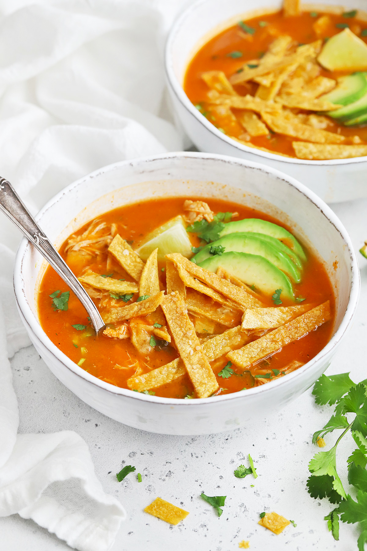 Front view of a white stoneware bowl of chicken tortilla soup topped with sliced avocado and crispy baked tortilla strips