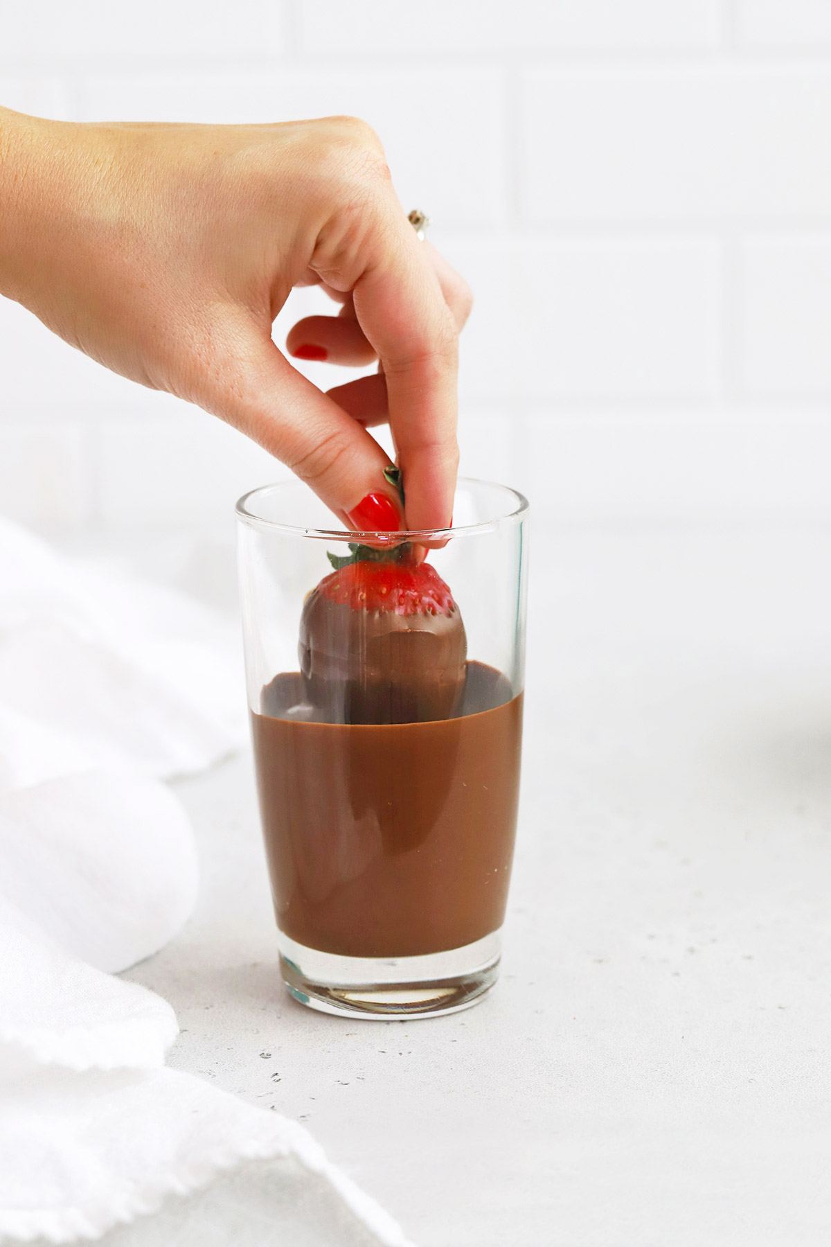 Front view of a hand dipping a strawberry into a glass of dairy-free chocolate